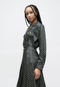 Vestido preto com padrão de laços dourados, mangas compridas, gola e dois bolsos no peito. Apresenta detalhes em botões dourados e cintura com cinto. Textura suave.