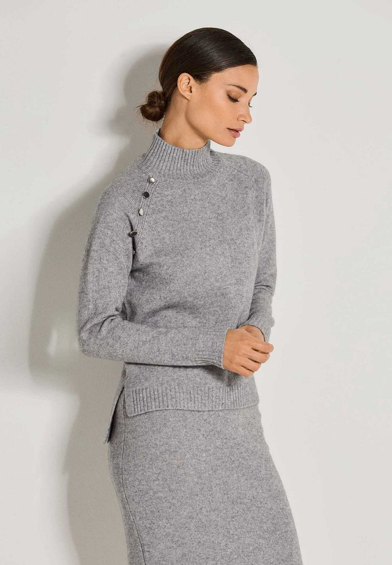 Frau mit dunklem Haar zu einem tiefen Dutt frisiert, trägt einen grauen Strickpullover mit Rollkragen und Knöpfen an der Schulter sowie einen passenden Rock.