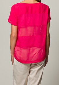 Fuksjowa bluzka z krótkim rękawem, z przezroczystym tyłem i jednolitym przodem; charakteryzuje się luźnym krojem i okrągłym dekoltem; lekki materiał.