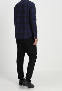 Camicia a quadri blu navy con maniche lunghe, abbinata a pantaloni neri a sigaretta e sneakers nere, vista da dietro.