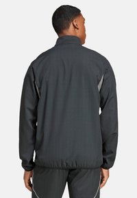 Schwarze Sportjacke mit hohem Kragen, langen Ärmeln und subtiler Satinoptik. Mit grauen Akzenten entlang der Seitennähte und Ärmelbündchen.