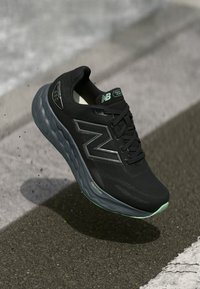 Melns New Balance skriešanas apavas ar pelēku zoli un zaļiem akcentiem, kas karājas virs asfalta ceļa netālu no baltās ielu līnijas.