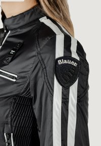 Giacca nera imbottita con strisce bianche, pannelli sulle spalle testurizzati, una tasca con zip e un patch circolare nero con il logo "Blauer".
