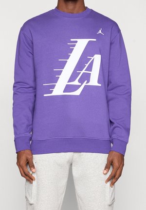 Mężczyzna noszący fioletową bluzę z dużym białym stylizowanym logo "LA" oraz małym logo Jumpman, w połączeniu z jasnoszarymi spodniami dresowymi.