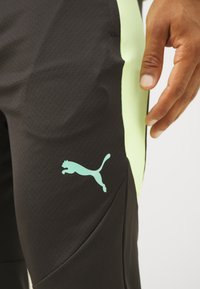 Schwarze Sporthosen mit strukturierter Oberfläche, ausgestattet mit neongrünen Seitenpanels und einem hellblauen Puma-Logo auf dem Oberschenkel.