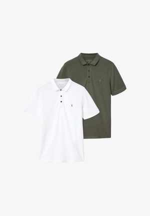 Mężczyzna w białej koszulce polo, czarnych szerokich spodniach i białych trampkach, trzymający czarną skórzaną torbę z dwoma przednimi kieszeniami, stojący na jednolitym tle.