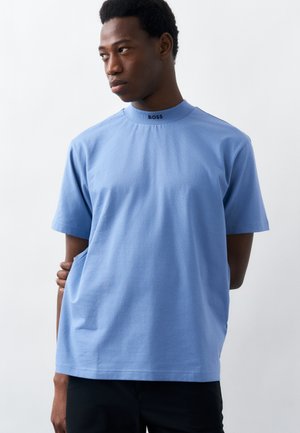 Νεαρός άνδρας με ανοιχτό μπλε κοντομάνικο πουκάμισο με το λογότυπο "BOSS" στο γιακά, στέκεται μπροστά σε απλό λευκό φόντο, κοιτώντας προς το πλάι.