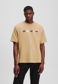 Beige pamut póló rövid ujjakkal és kerek nyakkivágással. Elöl négy fej ábrázolását tartalmazza, amelyeken napszemüveg van.