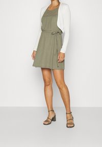 Robe vert olive avec un corsage smocké, une taille cintrée, et un cardigan court blanc. Associée à des talons gris à brides. Texture lisse.