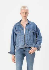 Giacca di jeans con una vestibilità rilassata, tonalità blu sbiadita, chiusura con bottoni, due tasche sul petto e maniche arrotolate per uno stile regolabile.
