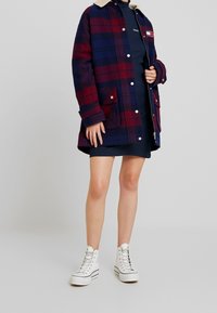 Granatowy i bordowy płaszcz w kratę z beżowym kołnierzem, bocznymi kieszeniami i zatrzaskami; noszony na granatowej sukience i zestawiony z białymi trampkami za kostkę.