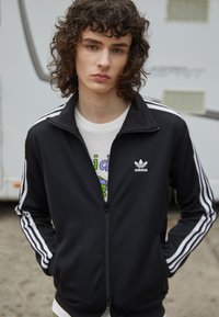 Svart treningsjakke med glidelås og hvite striper på ermene, kombinert med en hvit t-skjorte med grafisk trykk som har fargerik design.