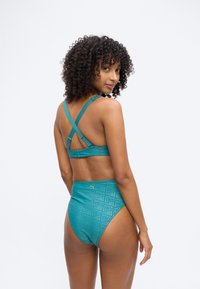 Νεαρή γυναίκα με σγουρά μαλλιά, που φοράει αθλητικό σουτιέν τιρκουάζ με σταυρωτούς ιμάντες και ψηλόμεσα εσώρουχα Puma, κοιτάζοντας πάνω από τον ώμο της σε λευκό φόντο.