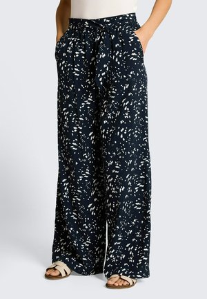 Granatowe szerokie spodnie z białymi abstrakcyjnymi plamami, z wiązanym pasem, boczne kieszenie, noszone z beżowymi sandałami i białym topem, pozą stojącą.