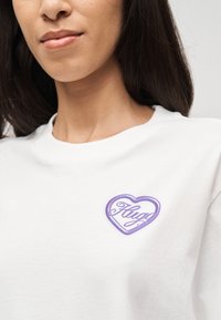 Κοντινό πλάνο γυναίκας που φοράει λευκό πουκάμισο με μοτίβο σε σχήμα μωβ καρδιάς και τη λέξη «Αγκαλιές» στο στήθος.