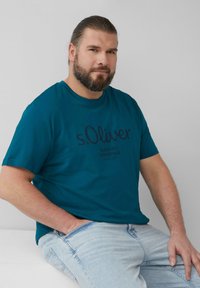 Teal katoenen T-shirt met korte mouwen, met een donkerblauw gedrukt logo en tekst. Gepaard met lichtblauwe denim jeans.