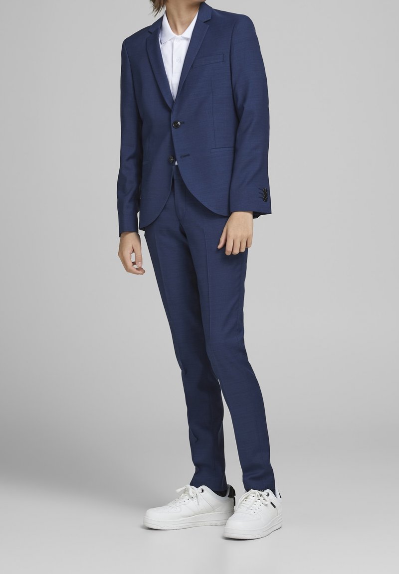 Personne portant un costume bleu marine ajusté avec une chemise blanche à col et des baskets blanches, debout devant un fond gris uni.