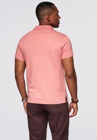 Růžová polo košile vyrobená z měkké látky, s krátkými rukávy a klasickým límcem. Má postranní rozparky a volný střih, doplněná tmavými kalhotami.