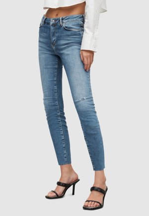 Jean slim bleu avec une légère déchirure au genou, porté avec des sandales noires à talons et une chemise blanche courte à manches longues.