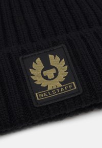 Svart stickad mössa med ribbad textur, prydd med en rektangulär patch med ett guld-emblem och texten "BELSTAFF".