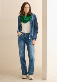 Veste en denim avec une surface texturée, T-shirt blanc, jeans bleu délavé et écharpe verte. Le modèle porte des baskets blanches pour un look décontracté.