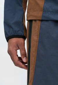 Ανδρικά αθλητικά εξωτερικά ρούχα με σχέδιο σε καφέ και μπλε χρώμα, ελαστικές μανσέτες, αντίθετες κατακόρυφες ρίγες και ορατή ραφή.