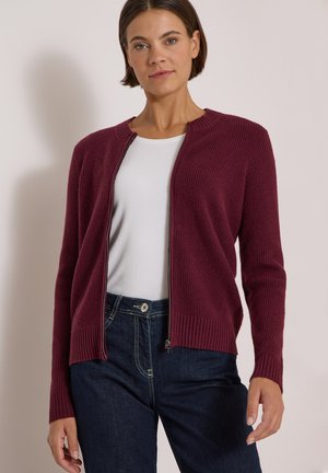 Vrouw die een bordeauxrode cardigan met rits draagt over een wit overhemd en donkerblauwe spijkerbroek, staand tegen een effen lichte achtergrond.