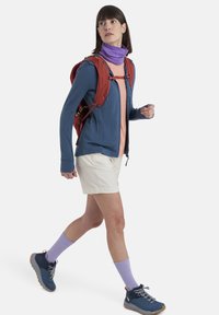 Giacca in pile blu, maglietta rosa chiaro, pantaloni corti crema, scaldacollo viola e calze, zaino rosso e scarpe sportive blu con dettagli arancioni.