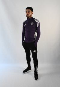 Top atlético púrpura con media cremallera y detalles en blanco, combinado con pantalones ajustados negros que cuentan con rayas y un logotipo en blanco, usados con zapatos negros.
