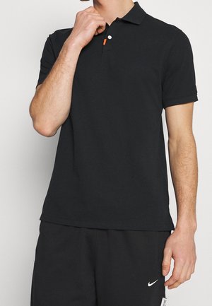 Man met een zwart polo-shirt met korte mouwen en zwarte Nike-shorts, met één hand bij de kraag en een neutrale achtergrond.