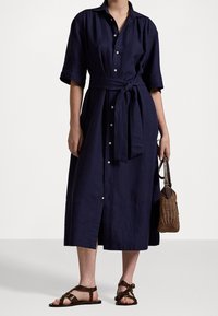 Mörkblå skjortklänning med korta ärmar, midjebälte och en texturerad tyg; matchad med bruna sandaler och en vävd väska.