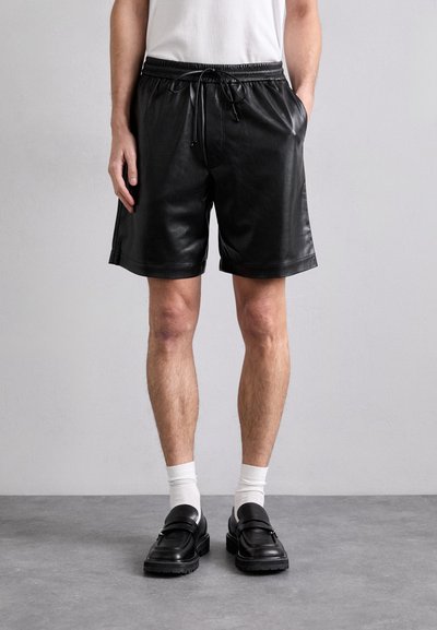 Sorte læder shorts med elastisk talje og snøre. Har en afslappet pasform og glat tekstur. Båret med sorte loafers og hvide sokker.