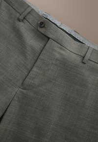 Pantalon de costume gris sur mesure avec ceinture boutonnée, passants et fermeture éclair cachée sur fond beige.