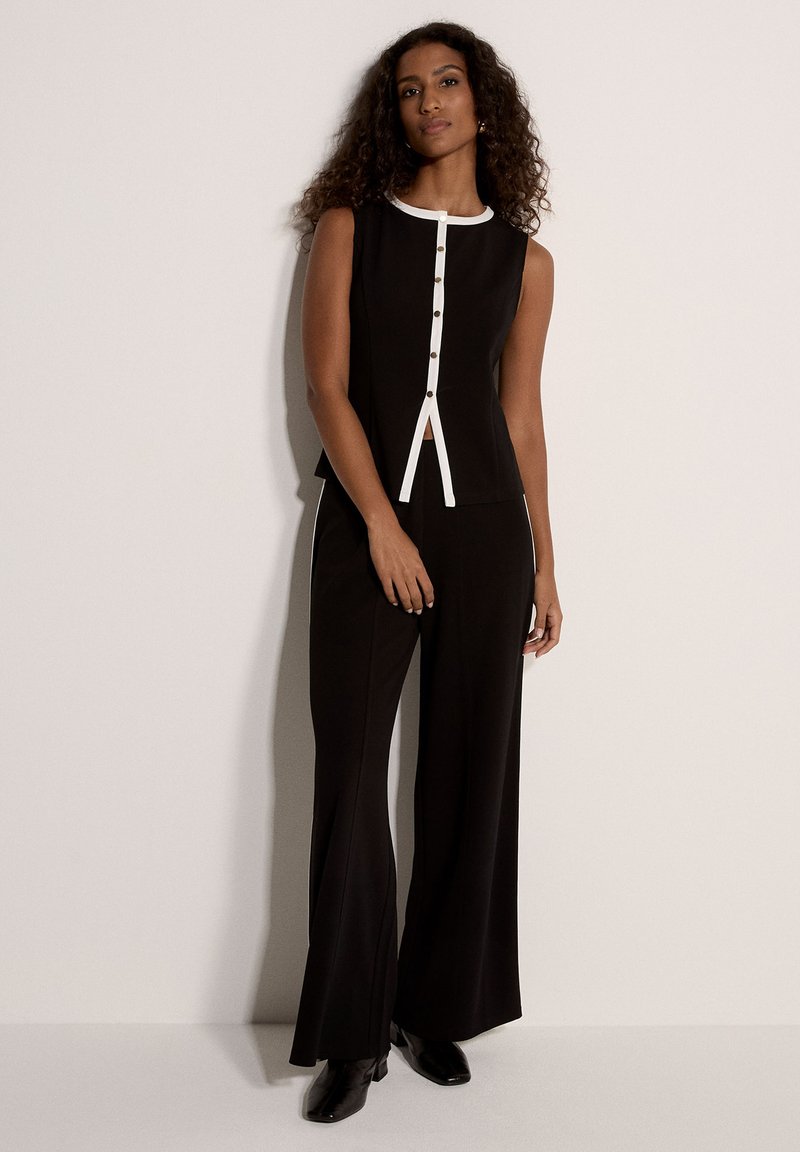 Haut noir sans manches avec bordure blanche et boutons, associé à un pantalon noir à jambes larges, présentant une silhouette épurée et structurée.