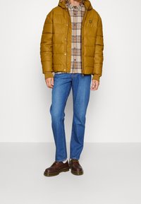 Hnědá prošívaná bunda s kapucí, kostkovaná košile, modré džíny a hnědé šněrovací boty. Bunda má malé logo na prsou.