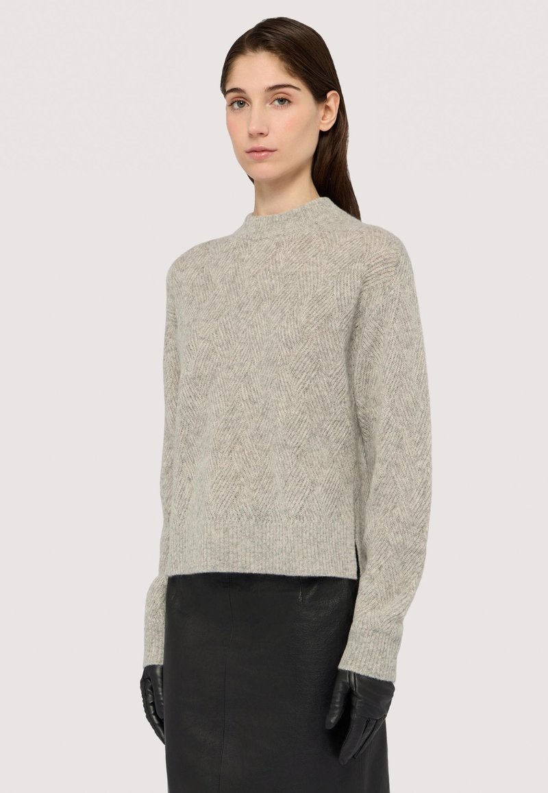 Hellgrauer, strukturierter Pullover mit Fischgrätmuster, hohem Kragen und überschnittenen Schultern, kombiniert mit Lederhandschuhen und einem schwarzen Rock.