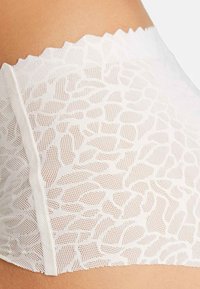 Sous-vêtements en dentelle blanche avec un motif floral portés sur la hanche d'une personne, montrant des détails en tissu en mailles fines et des bords festonnés.