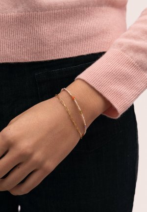 Muñeca con dos pulseras: una cadena delgada de oro y una delicada con pequeñas cuentas de colores, manga de suéter rosa, pantalones negros.