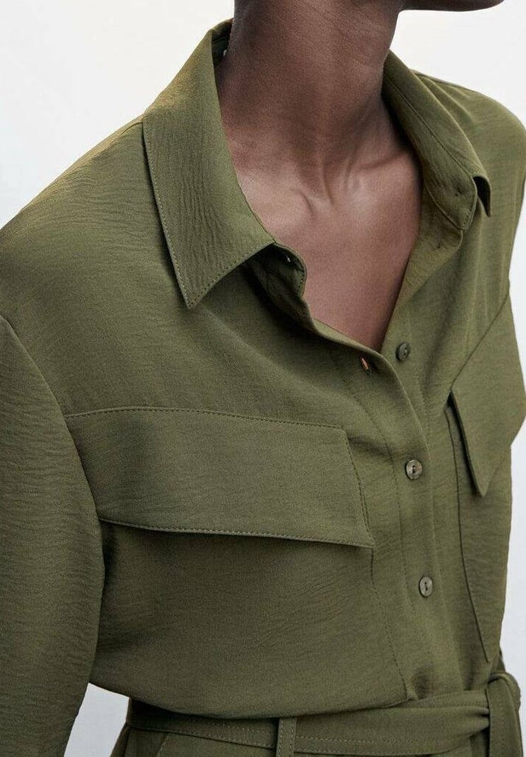 Gros plan d'une personne portant une chemise boutonnée vert olive avec de grandes poches poitrine et une ceinture à la taille.