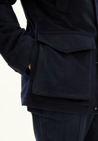 Tummansininen fleece-takki, jossa on suuri, litteä tasku ja pehmeä pinta. Takissa on tiukka istuvuus ja sivussa näkyvä napitus.