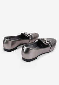 Mocasines gris metálico con suela plana, que cuentan con una hebilla metálica decorativa en el frente y detalles de costura a lo largo de los bordes.