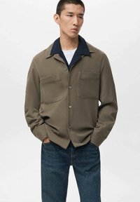 Camisa abotonada verde oliva con cuello azul marino, dos bolsillos en el pecho y un corte holgado, combinada con jeans de color azul oscuro. Textura de tela suave.