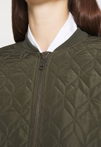 Giacca bomber trapuntata verde oliva con colletto a coste e chiusura con zip. Presenta cuciture a forma di diamante e una superficie liscia e texturizzata.