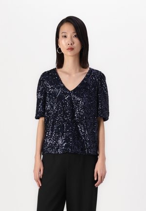 Marineblauw shirt met korte mouwen bedekt met pailletten, met een V-hals ontwerp. G gedragen met zwarte broek, die een losse pasvorm en een gestructureerd oppervlak heeft.