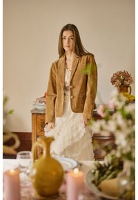 Jeune femme en blazer en daim marron et robe blanche en dentelle, debout près d'un buffet en bois avec des fleurs et des fruits dans une pièce doucement éclairée.