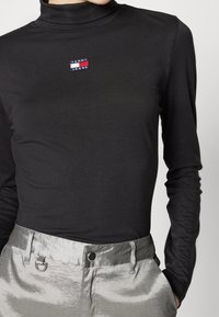 Svart långärmad polotröja med en liten röd, vit och blå rektangulär logotyp; matchad med ljusgrå, glänsande byxor.