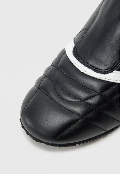 Μαύρο συνθετικό ποδοσφαιρικό παπούτσι με υφή καπιτονέ, στρογγυλεμένη μύτη και λευκή διακοσμητική ρίγα. Λαστιχένια σόλα με υφή για καλύτερη πρόσφυση.