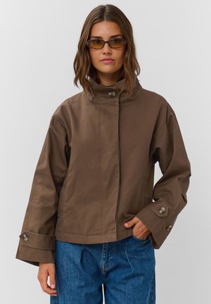 Femme portant une veste marron boutonnée avec de larges poignets et un jean bleu, une main dans la poche et des lunettes de soleil fines teintées marron.