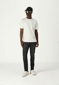 Mężczyzna w białej koszulce, czarnych dżinsach, białych trampkach, czapce w kolorze czarnym i białych okularach przeciwsłonecznych stoi na tle czystej białej ściany.