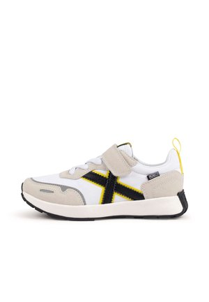 Baskets avec une tige en tissu blanc, accents noirs et jaunes, surpiqûres en daim et une bride à scratch. Caractérisées par une semelle en caoutchouc texturée et une languette de tirage.
